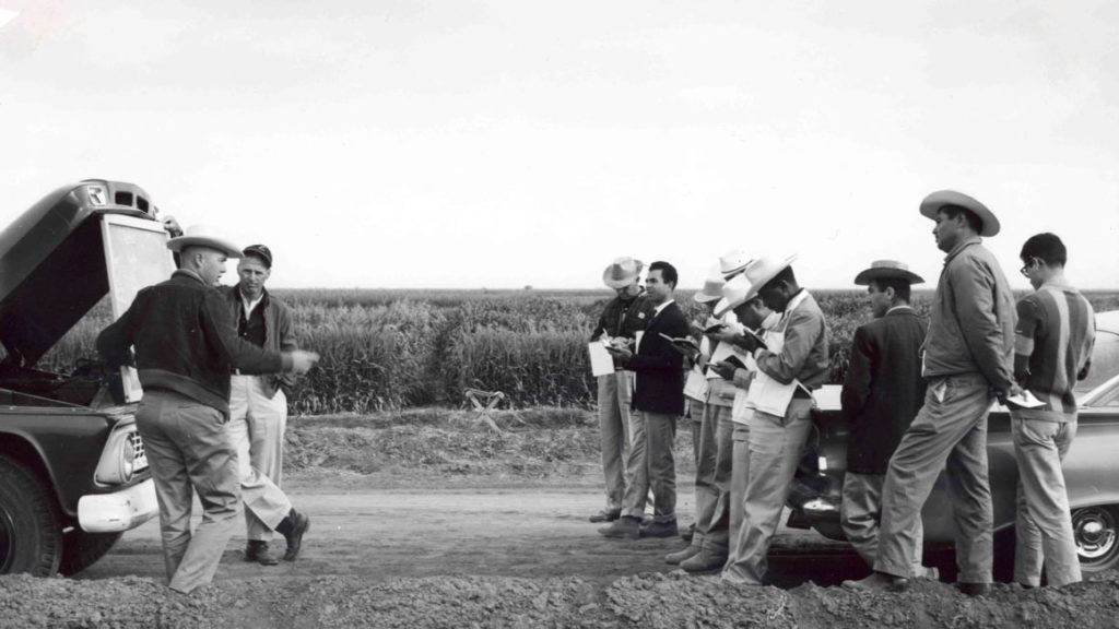 Black and white image of a group of men standing in front of vechicles. Men on the left side of the image stand in front of a truck with a chalk board speaking towards the men on the right side of the photo. Men on the right side, standing in front of a car, are taking notes.