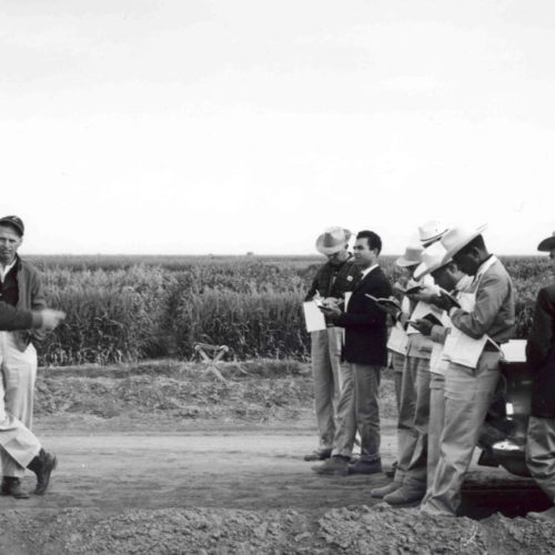 Black and white image of a group of men standing in front of vechicles. Men on the left side of the image stand in front of a truck with a chalk board speaking towards the men on the right side of the photo. Men on the right side, standing in front of a car, are taking notes.