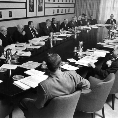 Two dozen men sit around a long oval table, with stacks of paper in front of each one. Some of the men are smoking.