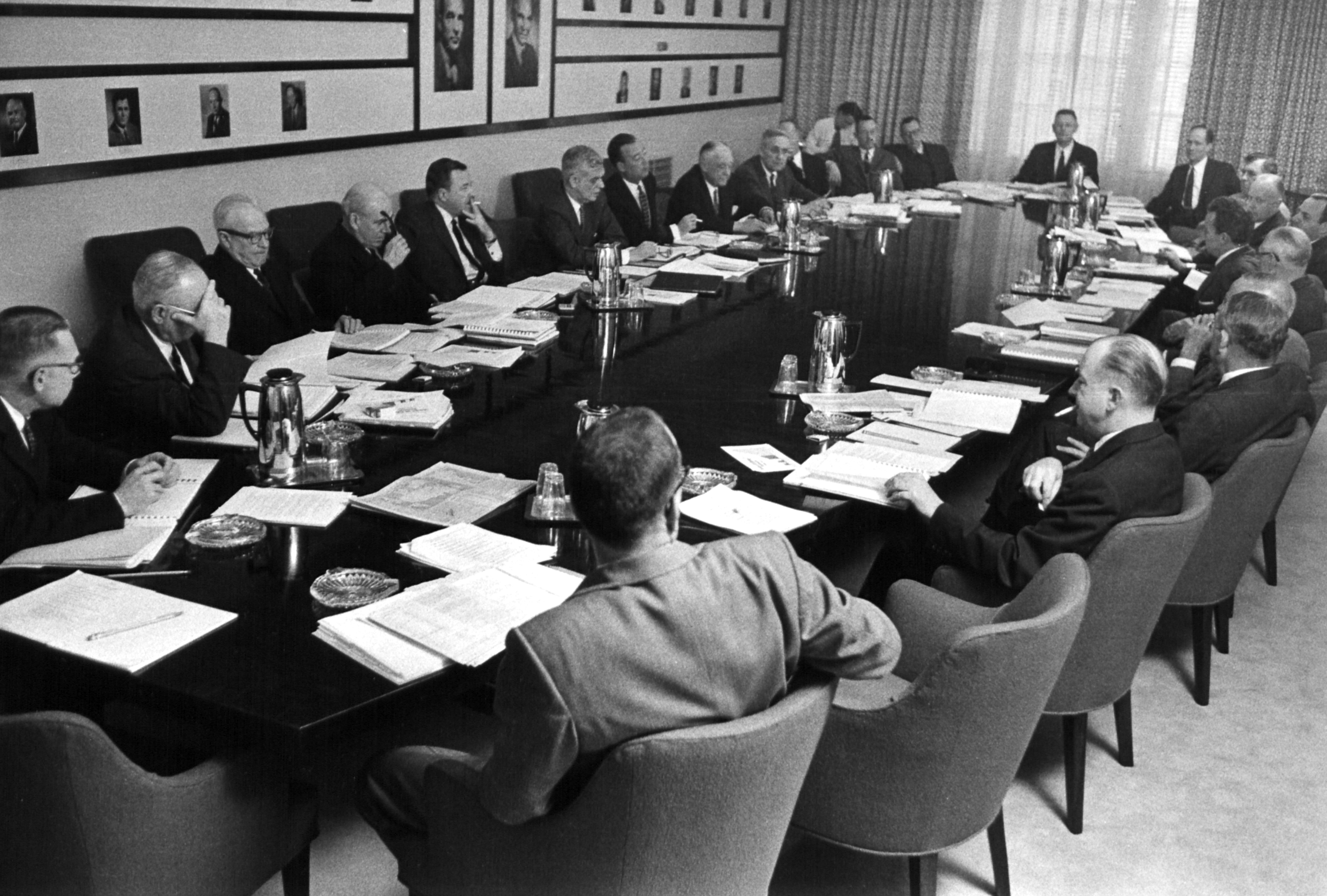 Two dozen men sit around a long oval table, with stacks of paper in front of each one. Some of the men are smoking.