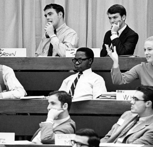 Nine students listen to a lecture in a lecture hall. A female student raises her hand.