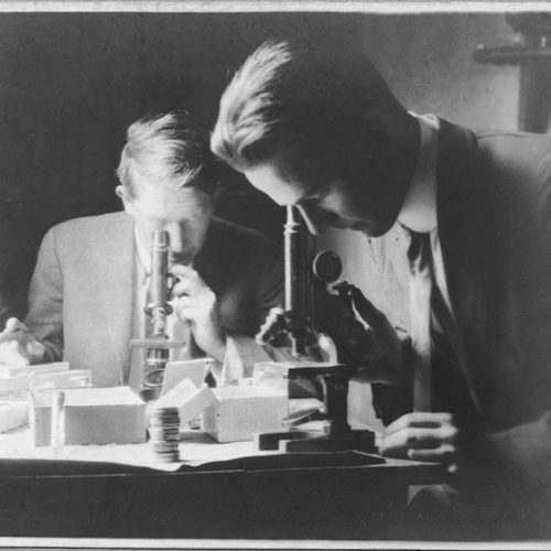 Black and white image of two scientist using microscopes surrounded by other scientific equipment.